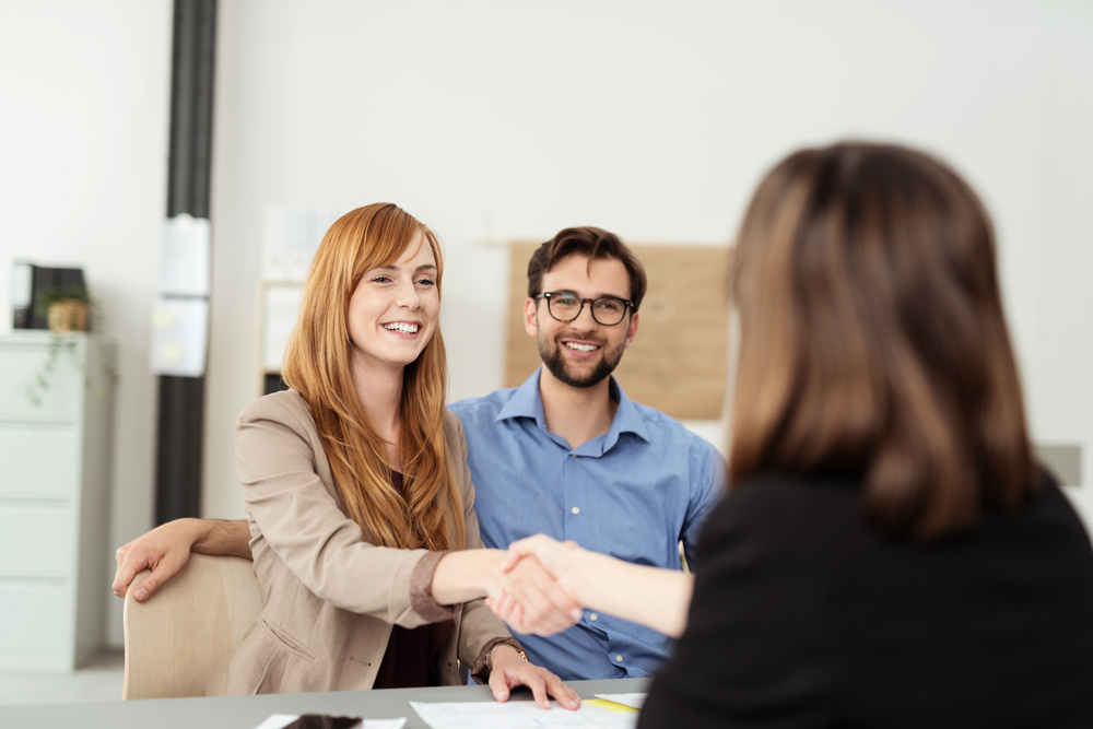 Happy young couple meeting with a broker in her office leaning over the desk to shake hands, view from behind the female agent-2